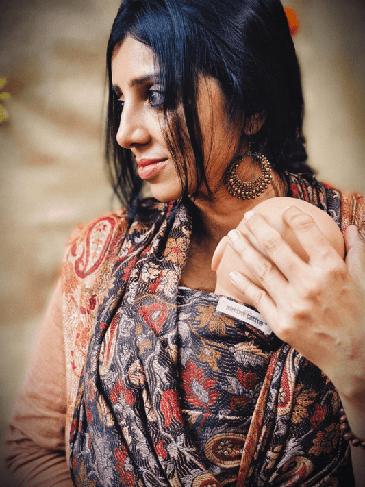 Caregiver wearing a cashmere babywearing wrap in autumnal hues, holding a newborn in a high, ergonomic front carry. The woven wrap sling provides adjustable support, skin-to-skin closeness, and a secure fit for inclusive caregivers.