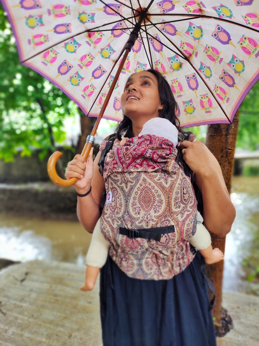 Front-facing view of Ishkabon Yoga newborn baby carrier in earthy paisley print, adjustable ergonomic carrier with soft merino blend fabric, baby rests chest-to-chest, modern hands-free parenting lifestyle