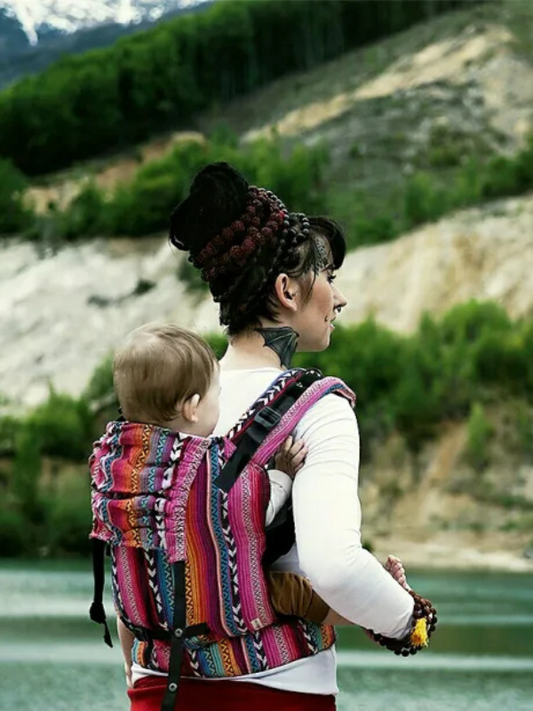 Caregiver wearing Larona Nomad ergonomic baby carrier in vibrant woven pink and multicolor fabric, back carry position with toddler, breathable structured baby carrier designed for hands-free parenting, adventure-ready comfort, and inclusive babywearing for all caregivers.