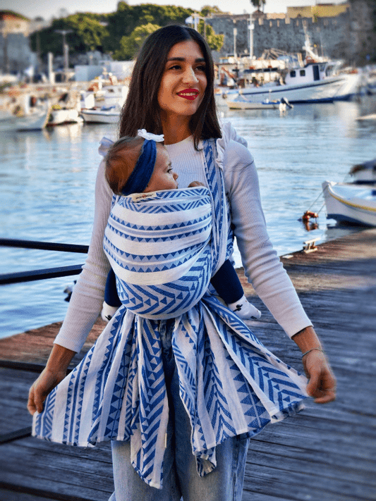 Caregiver wearing Neelanjana blue and white woven wrap baby carrier while walking on a dock by the harbor, front carrying baby in ergonomic with flowing wrap tails, breathable cotton newborn wrap for hands-free parenting and everyday movement.