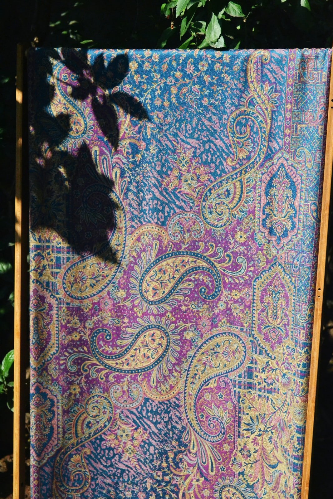 Full-length view of a woven marisa baby wrap by Almitra Tattva displayed outdoors, featuring intricate paisley and floral motifs in teal, violet, gold, and soft pink tones. The breathable woven baby carrier fabric highlighting texture, pattern density, and artisanal craftsmanship. Designed as an ergonomic baby wrap sling for newborns and toddlers, suitable for hands-free parenting and all caregivers.