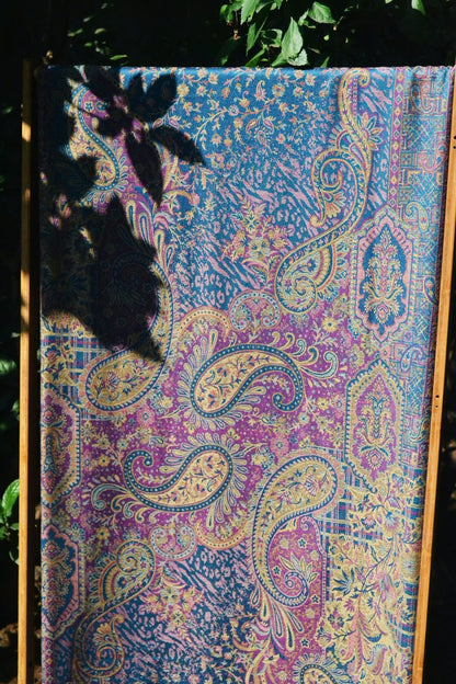 Full-length view of a woven marisa baby wrap by Almitra Tattva displayed outdoors, featuring intricate paisley and floral motifs in teal, violet, gold, and soft pink tones. The breathable woven baby carrier fabric highlighting texture, pattern density, and artisanal craftsmanship. Designed as an ergonomic baby wrap sling for newborns and toddlers, suitable for hands-free parenting and all caregivers.