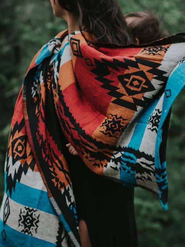 Caregiver wearing the Auronee ergonomic baby wrap carrier in an outdoor setting, featuring a bold handwoven tribal pattern in rust, black, teal, and ivory tones, supporting a secure front carry for newborns and babies with breathable fabric and hands-free babywearing comfort for all caregivers.