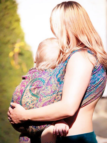 Caregiver carrying a baby in an ergonomic wrap using a colorful. The newborn wrap sling supports a secure front carry with even weight distribution, allowing hands-free movement outdoors. Gender-neutral babywearing and everyday conscious parenting.