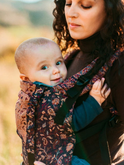 Smiling baby nestled in the Sapphire Yoga Baby Carrier worn by a parent outdoors, showcasing ergonomic design and traditional weave.