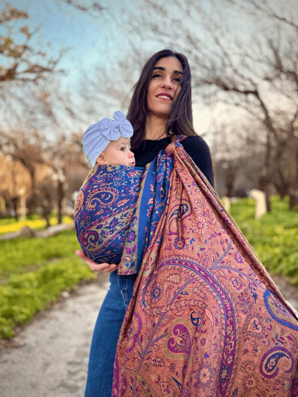 Close-up view of a newborn wrap carrier in rich blue and rust paisley fabric, showing snug ergonomic support for baby’s back and hips while worn by a caregiver outdoors, designed for breathable comfort and hands-free parenting.