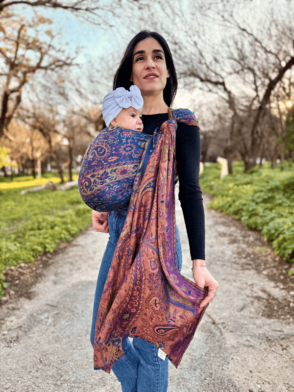 Caregiver wearing a Falak Woven Wrap ergonomic baby sling in deep blue and copper tones, carrying an infant hands-free in a front carry position outdoors, showcasing breathable fabric and secure newborn support.