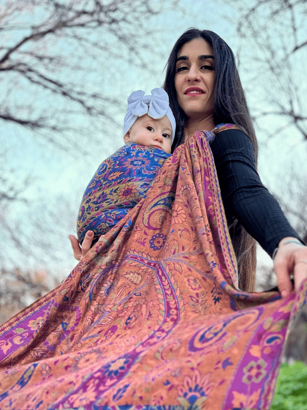 Low-angle view of a flowing paisley baby wrap sling draped across the caregiver’s body, demonstrating fabric length, soft structure, and ergonomic newborn carry design in a natural outdoor setting.