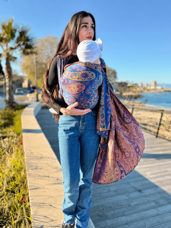 Caregiver walking along a seaside boardwalk using a falak wrap wrap sling baby carrier, featuring breathable woven fabric, front carry positioning, and hands-free support for infants in outdoor everyday use.
