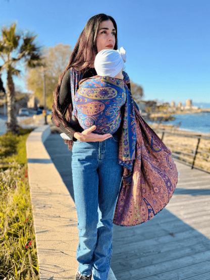 Caregiver walking along a seaside boardwalk using a falak wrap wrap sling baby carrier, featuring breathable woven fabric, front carry positioning, and hands-free support for infants in outdoor everyday use.