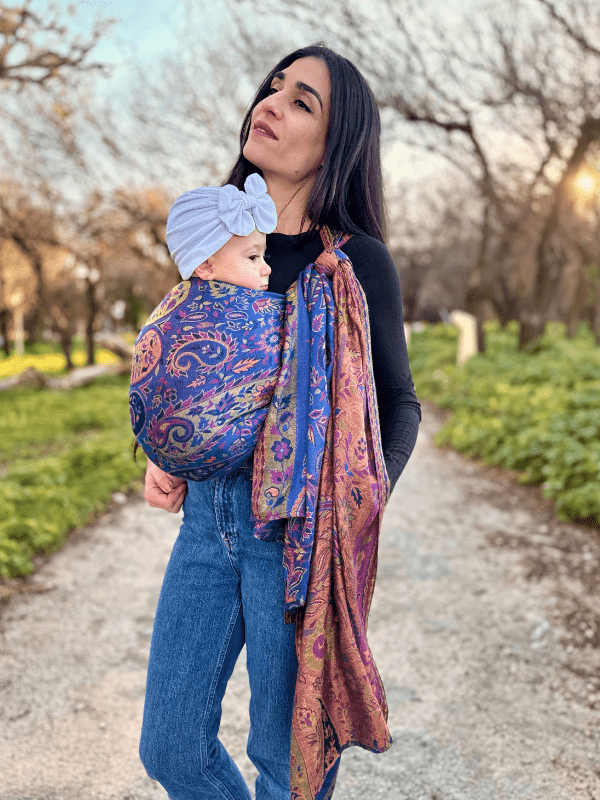 Caregiver wearing a front carry baby wrap carrier while standing on a park path, showing secure ergonomic positioning, hands-free comfort, and inclusive babywearing design for everyday movement.