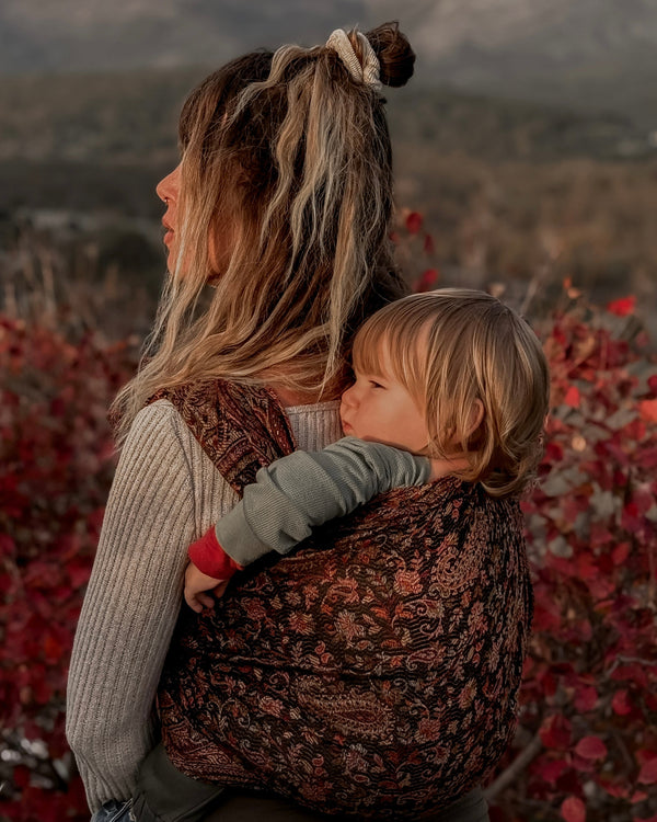 Woman carrying a child in a floral baby carrier with a mountainous landscape in the background