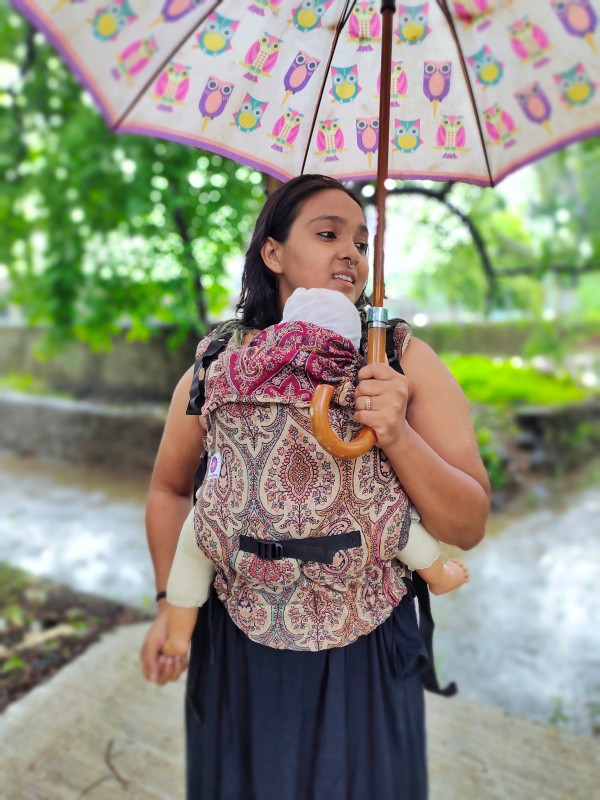 Caregiver wearing Ishkabon Yoga ergonomic baby carrier in paisley merino blend fabric, front carrying newborn outdoors under an umbrella, breathable structured carrier with padded straps for hands-free parenting in nature