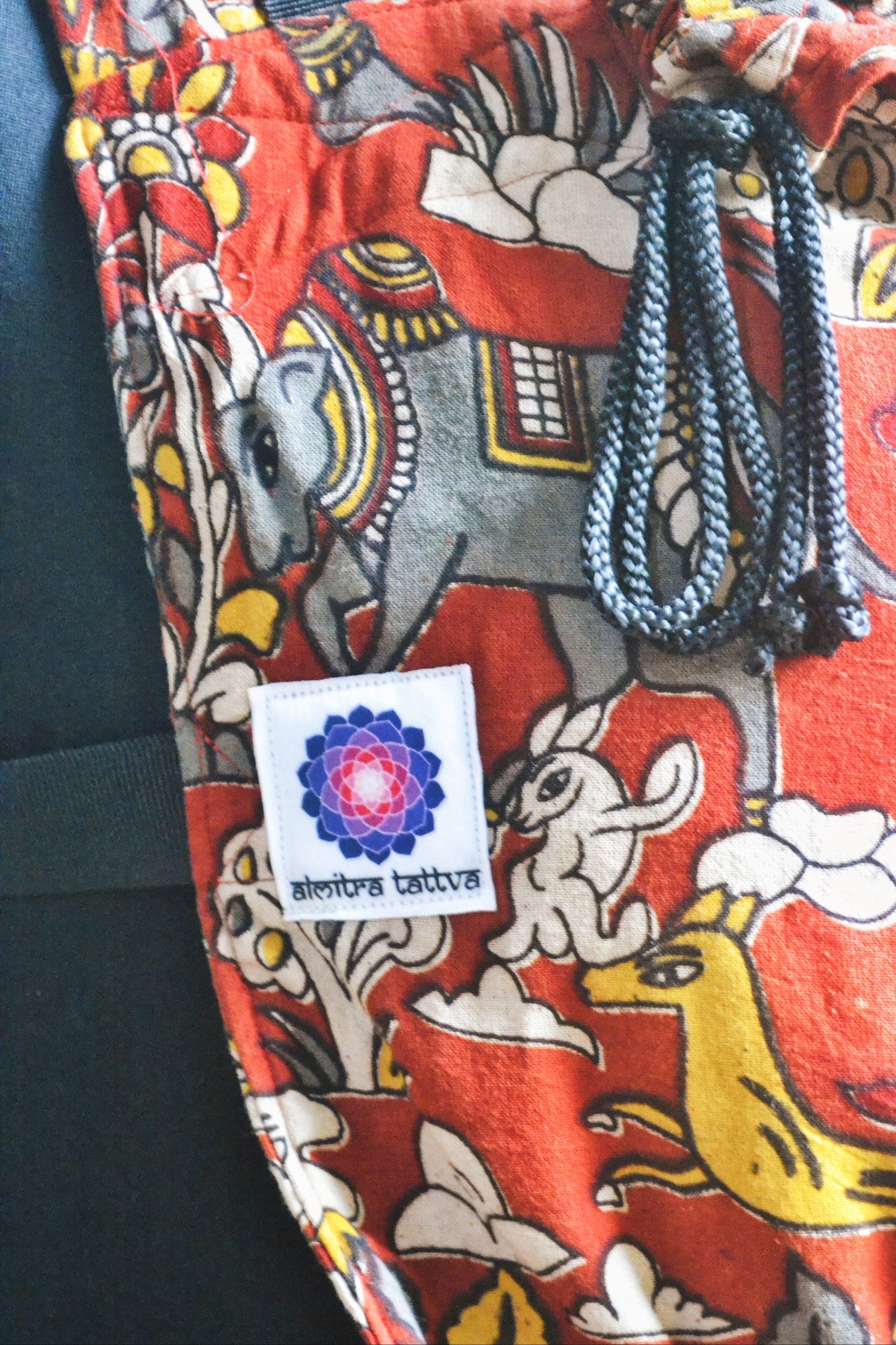 Jataka Tales Kalamkari baby carrier fabric showing Almitra Tattva logo label, with animal and folk motifs, breathable soft structured newborn carrier with drawstring hood detail for ergonomic babywearing.