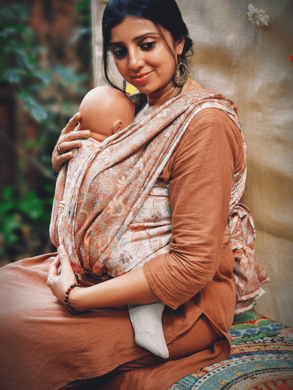 Caregiver seated while wearing an ergonomic baby wrap carrier in muted terracotta hues. The woven wrap sling supports a calm, secure newborn carry with breathable fabric and adaptive comfort for extended wear.