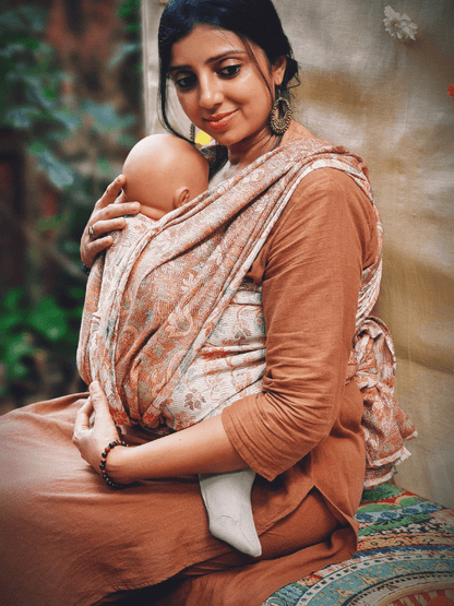 Caregiver seated while wearing an ergonomic baby wrap carrier in muted terracotta hues. The woven wrap sling supports a calm, secure newborn carry with breathable fabric and adaptive comfort for extended wear.