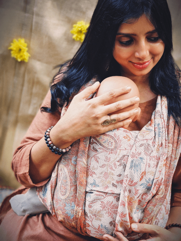 Detail view of a newborn baby nestled in a soft ergonomic wrap carrier with warm neutral tones. The wrap sling provides full body support, even weight distribution, and a secure fit for inclusive babywearing across caregivers.