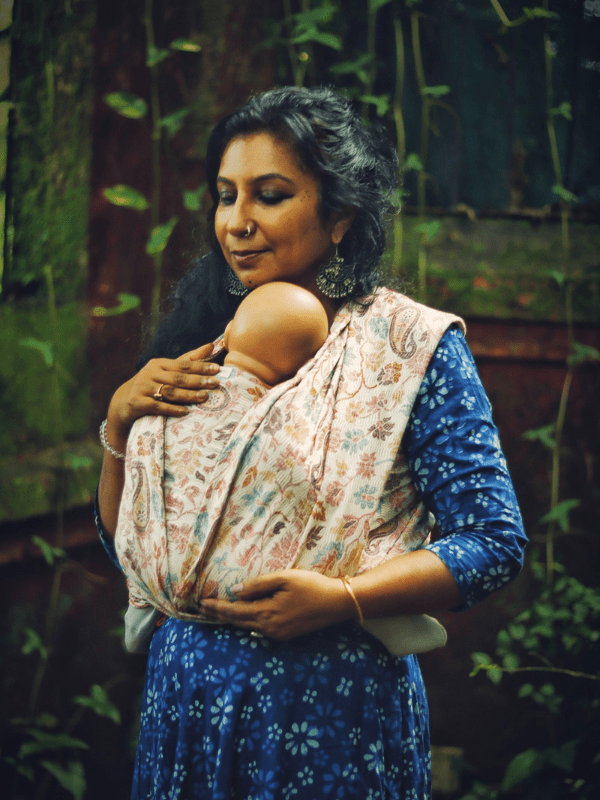 Caregiver wearing a soft woven baby wrap sling in natural tones, holding a baby in a front carry position outdoors. The ergonomic baby carrier offers breathable comfort, grounded aesthetics, and hands-free parenting for all caregivers.