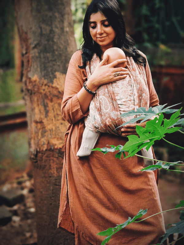 Caregiver wearing a terracotta-toned ergonomic newborn wrap sling, holding a sleeping baby close to the chest. Soft woven fabric supports hands-free babywearing in a natural outdoor setting, designed for newborn comfort and mindful parenting.