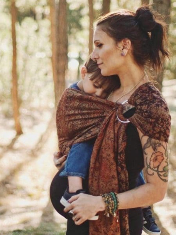Mother babywearing toddler in Almitra Tattva Kalindi Ring Sling, showing ergonomic support, rust-earth tones, and broad shoulder spread in forest setting.