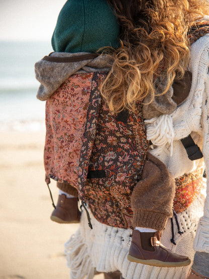 Close-up back carry view of Kalindi Cashmere ergonomic baby carrier in rust paisley floral jamawar wool, toddler seated in breathable structured carrier on caregiver’s back, supportive fit for hands-free parenting, premium woven fabric designed for conscious, inclusive babywearing.