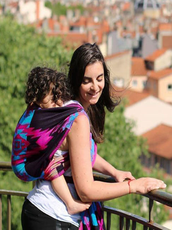 Caregiver wearing the Kullu Collection ergonomic baby wrap carrier in a secure back carry, featuring bold geometric patterns in teal, magenta, and black. Soft breathable woven fabric supports a toddler in an ergonomic position while enabling hands-free parenting in an urban outdoor setting. 