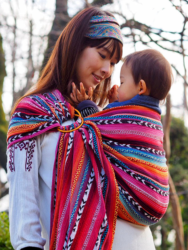 Mother babywearing in Almitra Tattva’s Larona Ring Sling – vibrant rainbow handwoven baby carrier from the Bohemian Collection, worn outdoors in winter.