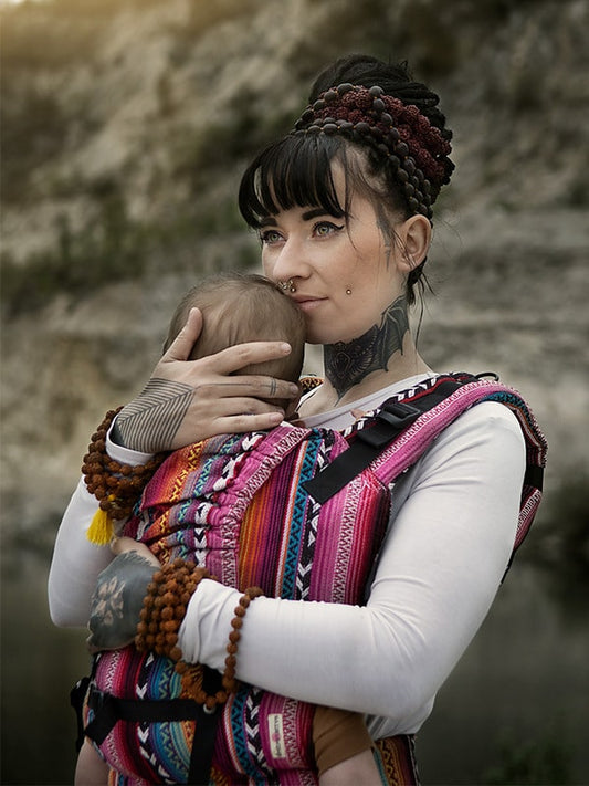 Holding baby in Larona soft structured baby carrier, front carry position with bold pink woven textile, ergonomic newborn carrier supporting natural posture, breathable fabric for long wear, secure fit for conscious, gender-fluid babywearing in outdoor lifestyle setting.