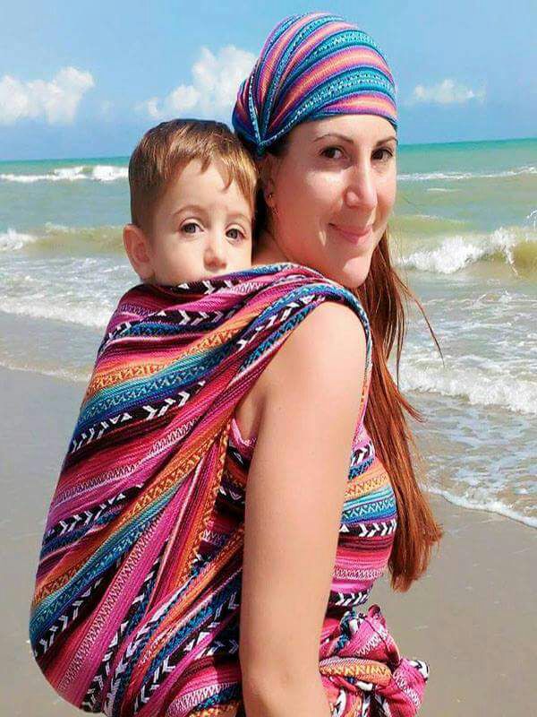Caregiver wearing a Larona Woven Wrap Carrier ergonomic at the beach, carrying a baby in a secure back carry position, made from breathable handwoven fabric, supporting newborn to toddler babywearing, hands-free parenting, and inclusive, gender-fluid outdoor lifestyle use.