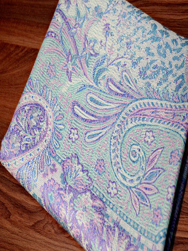 Folded pastel Jamawar baby wrap sling resting on a wooden surface, showcasing soft wool-blend fabric, detailed paisley motifs, and a compact fold ideal for everyday ergonomic baby carrying and on-the-go conscious parenting.