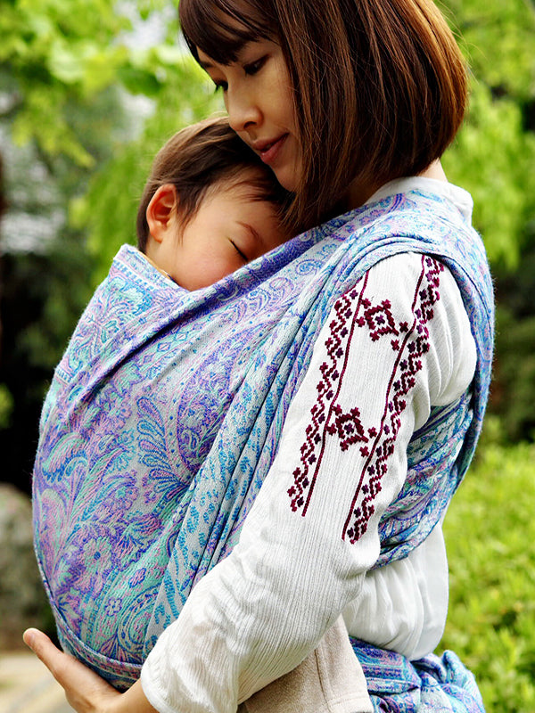 Angled close-up of Marisa White baby carrier fabric highlighting flowing paisley motifs in pastel aqua, lavender, and mint, demonstrating soft structure, breathable weave, and supportive design for ergonomic hands-free babywearing.