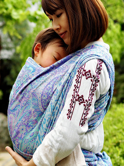 Angled close-up of Marisa White baby carrier fabric highlighting flowing paisley motifs in pastel aqua, lavender, and mint, demonstrating soft structure, breathable weave, and supportive design for ergonomic hands-free babywearing.