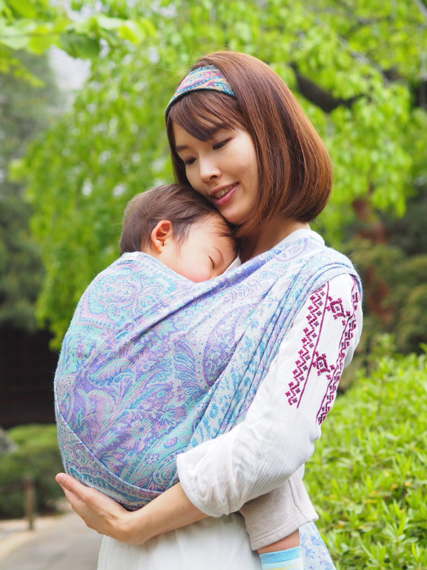 Woman holding a baby wrapped in a Marisa White Woven Wrap - Cashmere Collection outdoors with greenery in the background