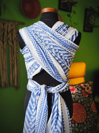 Neelanjana blue and white woven baby wrap, showing crisscross carry with long tail tie, breathable cotton fabric and geometric pattern, designed for ergonomic babywearing, hands-free parenting, and inclusive caregivers.