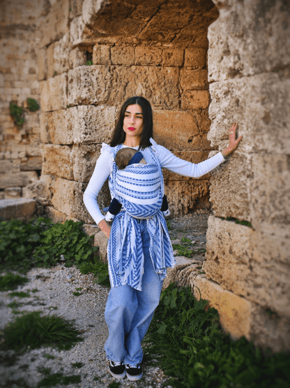 Caregiver wearing Neelanjana blue and white woven baby carrier outdoors with baby in front carry position, featuring wrap-style tails, breathable cotton fabric, and ergonomic support for inclusive, hands-free parenting in everyday lifestyle settings.