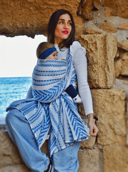 Caregiver babywearing with Neelanjana ergonomic baby wrap, baby held in front carry, blue and white woven cotton fabric with wrap-around ties, designed for newborn support, breathable comfort, and bold gender-fluid hands-free parenting.