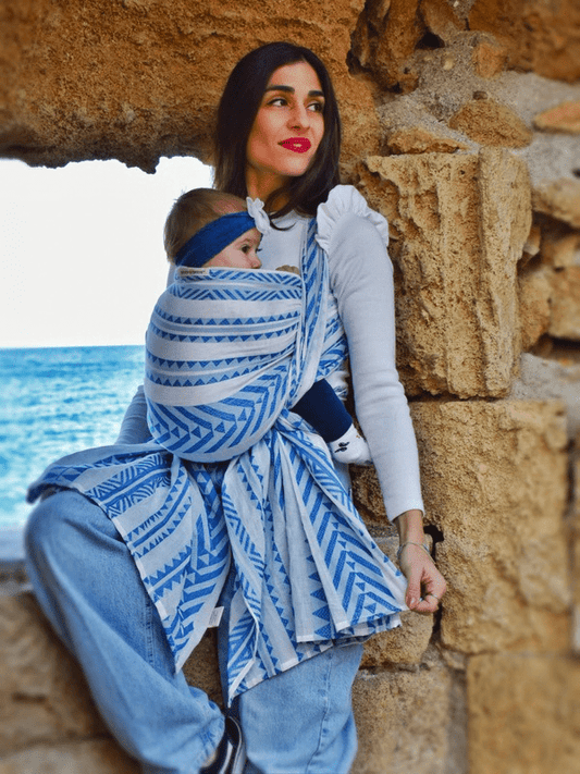 Caregiver babywearing with Neelanjana ergonomic baby wrap, baby held in front carry, blue and white woven cotton fabric with wrap-around ties, designed for newborn support, breathable comfort, and bold gender-fluid hands-free parenting.