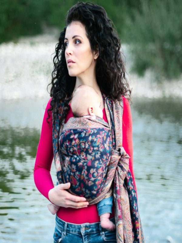 Caregiver wearing a front carry  newborn in nilofer wrap sling made from breathable silk blend fabric with rich paisley detailing, offering snug ergonomic support for baby while allowing comfortable movement during outdoor babywearing.
