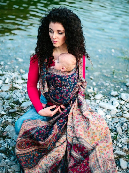 Seated caregiver using a woven ergonomic baby wrap with flowing borders and floral motifs, supporting a newborn securely against the chest, emphasizing fabric drape, comfort, and inclusive hands-free parenting design. 