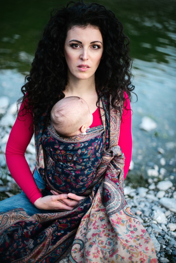 Front carry Nilofer babywearing setup using a woven paisley baby wrap in navy and muted florals, supporting a newborn in an ergonomic, worn by a caregiver seated near water, highlighting softness, drape, and secure wrap sling support.