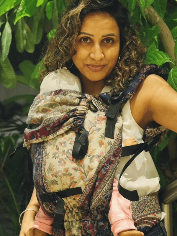 Nilofer wrap-inspired soft structured baby carrier in floral wool, newborn supported in ergonomic front carry with breathable textile and adjustable shoulder straps, designed for inclusive hands-free babywearing.