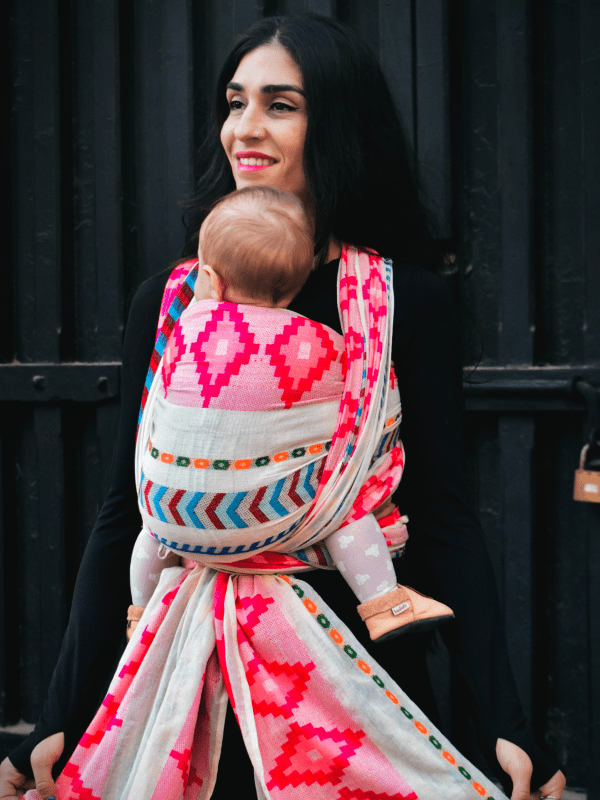 Front-facing view of Shubra ergonomic baby wrap worn by caregiver during an outdoor stroll, featuring bold geometric patterns, wide shoulder support, and secure newborn carry in a woven baby carrier.