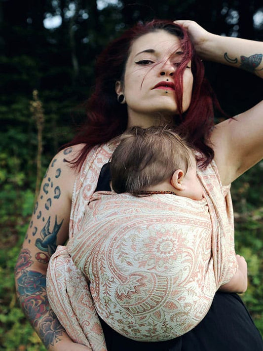 Caregiver wearing the Tushara baby wrap carrier outdoors, carrying a baby in a secure front carry position, featuring a softly structured woven fabric in warm neutral tones, supporting ergonomic babywearing and hands-free movement for modern caregivers.