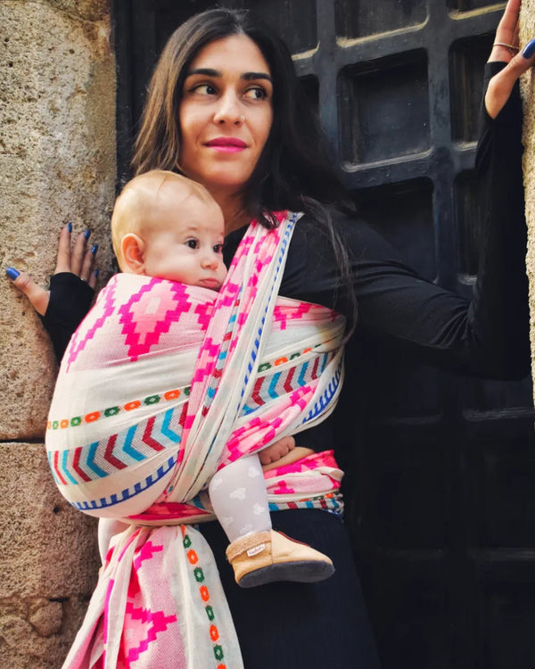 Woman holding a baby in a colorful sling against a stone wall.