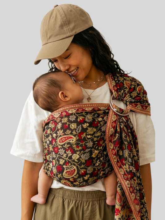 Smiling mother in a tan cap gently touching her baby's cheek while carrying them in a dark red and gold floral paisley Almitra Tattva Chenab 2.0 ring sling.