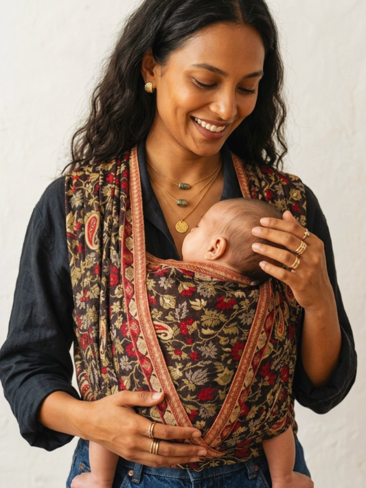 A woman wearing a Ababy carrier wrap with a sleeping baby inside, photographed against a plain light wall.