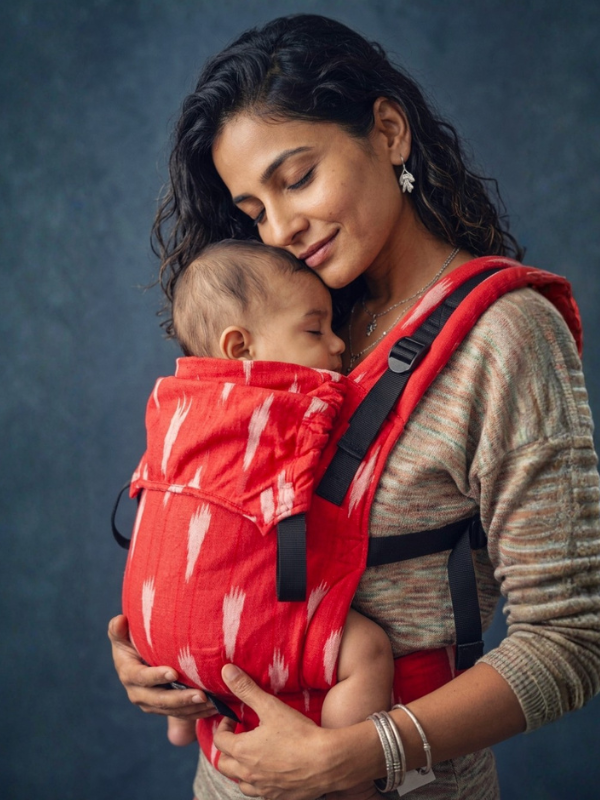 A woman wearing a red baby carrier with a white ikat pattern holds a sleeping baby against her chest. The baby carrier has black adjustable straps and is shown in a studio setting with a dark textured blue-gray background.