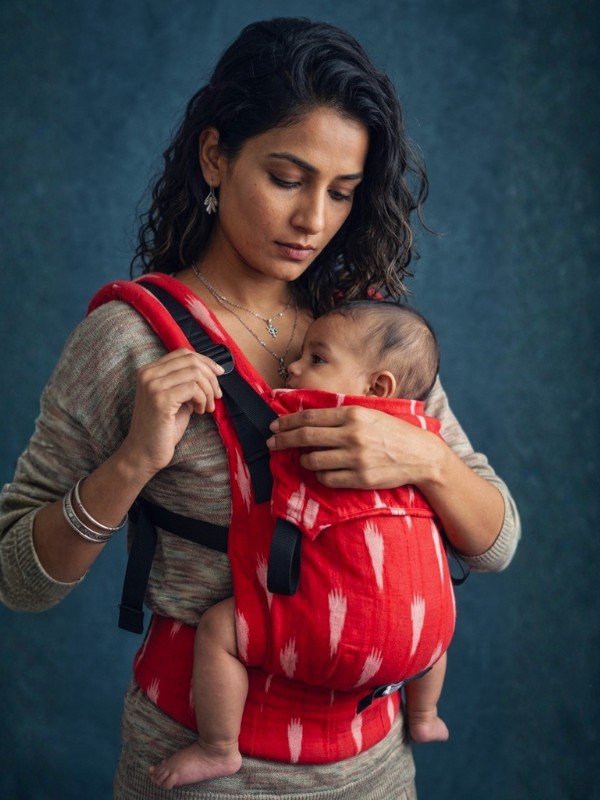 A baby carrier worn on a person's back, shown from behind. The carrier has green and black patterned fabric panels with black adjustable straps and buckles.