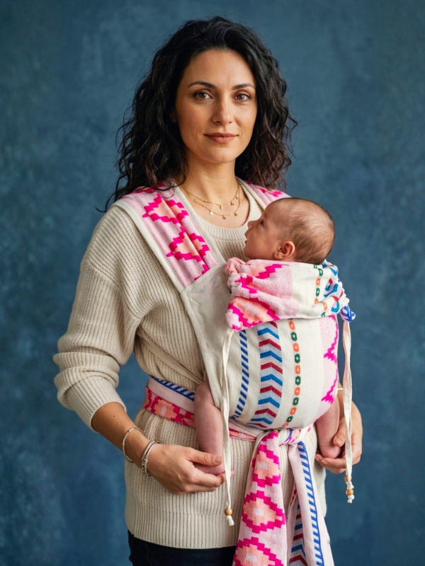 A woman wearing a cream knit sweater and dark pants carries a baby in a pink, white, and blue patterned wrap carrier against a textured blue studio background.