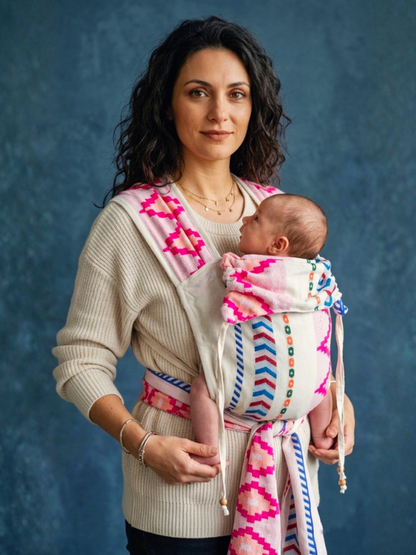 A woman wearing a cream knit sweater and dark pants carries a baby in a pink, white, and blue patterned wrap carrier against a textured blue studio background.